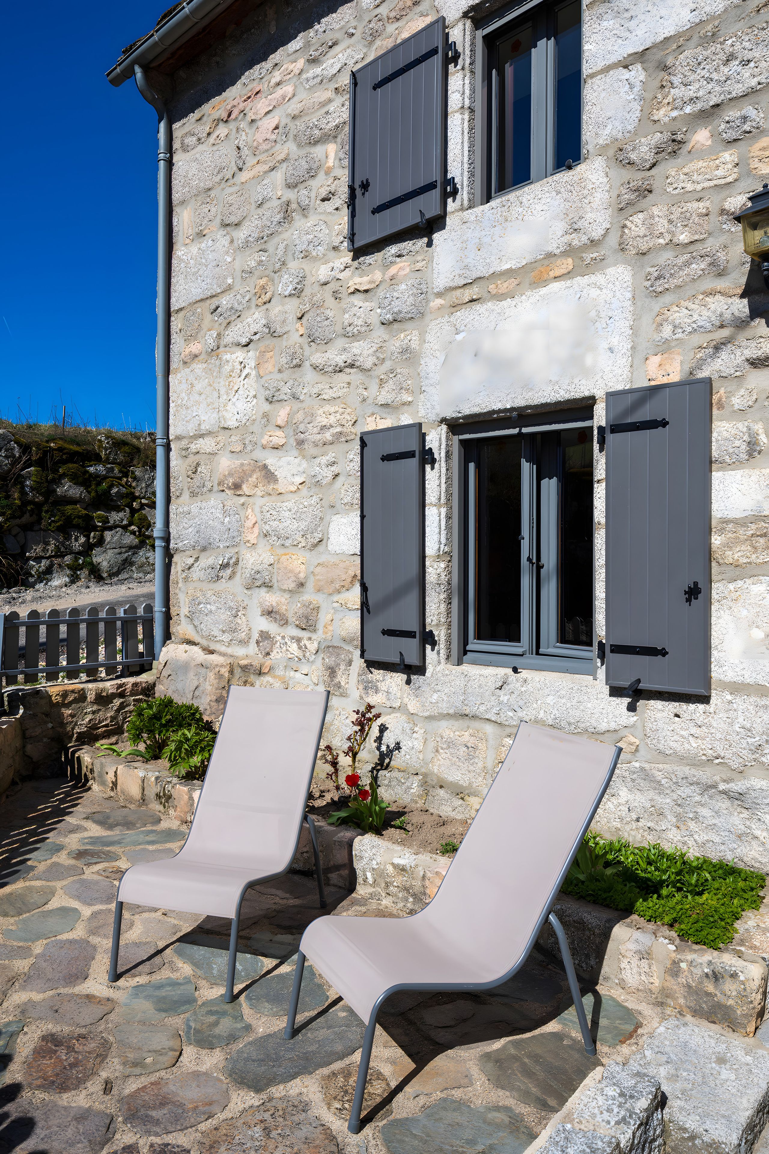 Un edificio de piedra con dos ventanas y contraventanas, un área pavimentada con dos sillones de descanso y un pequeño jardín con flores rojas.