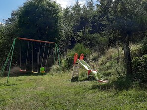 A playground with swings and a slide in a grassy area surrounded by trees.