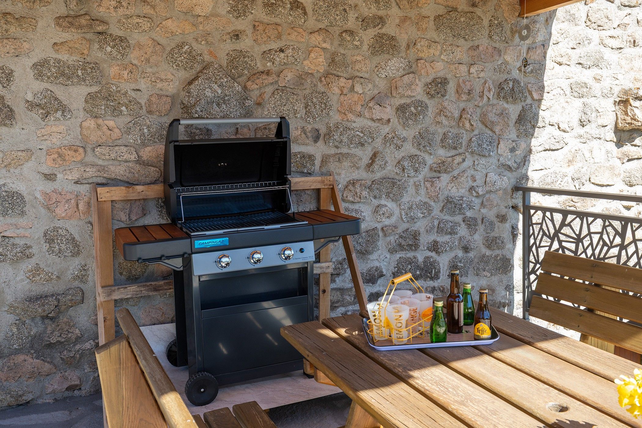 Un barbecue portable doté d’une table et de bancs en bois, installé devant un mur de pierres.