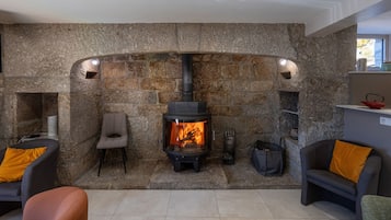 En stenkamin med en vedeldad kamin, en sittgrupp med stolar och en liten hylla med diverse föremål.