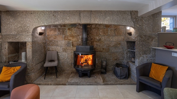 A stone fireplace with a wood-burning stove, seating area with chairs, and a small shelf with items.