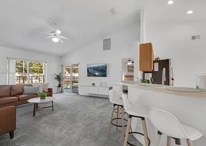 Ein modernes Wohnzimmer mit Sofa, Couchtisch und Sesseln, ein Küchenbereich mit Bar und ein großes Fenster mit Blick.