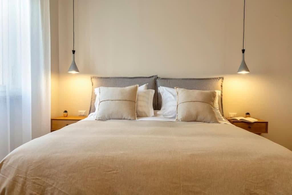 Un letto rifatto con cura, con una testiera grigia, due comodini con lampade e una finestra con tende.