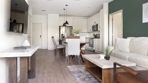 A modern kitchen with white cabinets, a dining area with a wooden table and chairs, and a living room with a sofa and a coffee table. - Landing Amazing 1bd, Gym, Game Room (Gainesville)