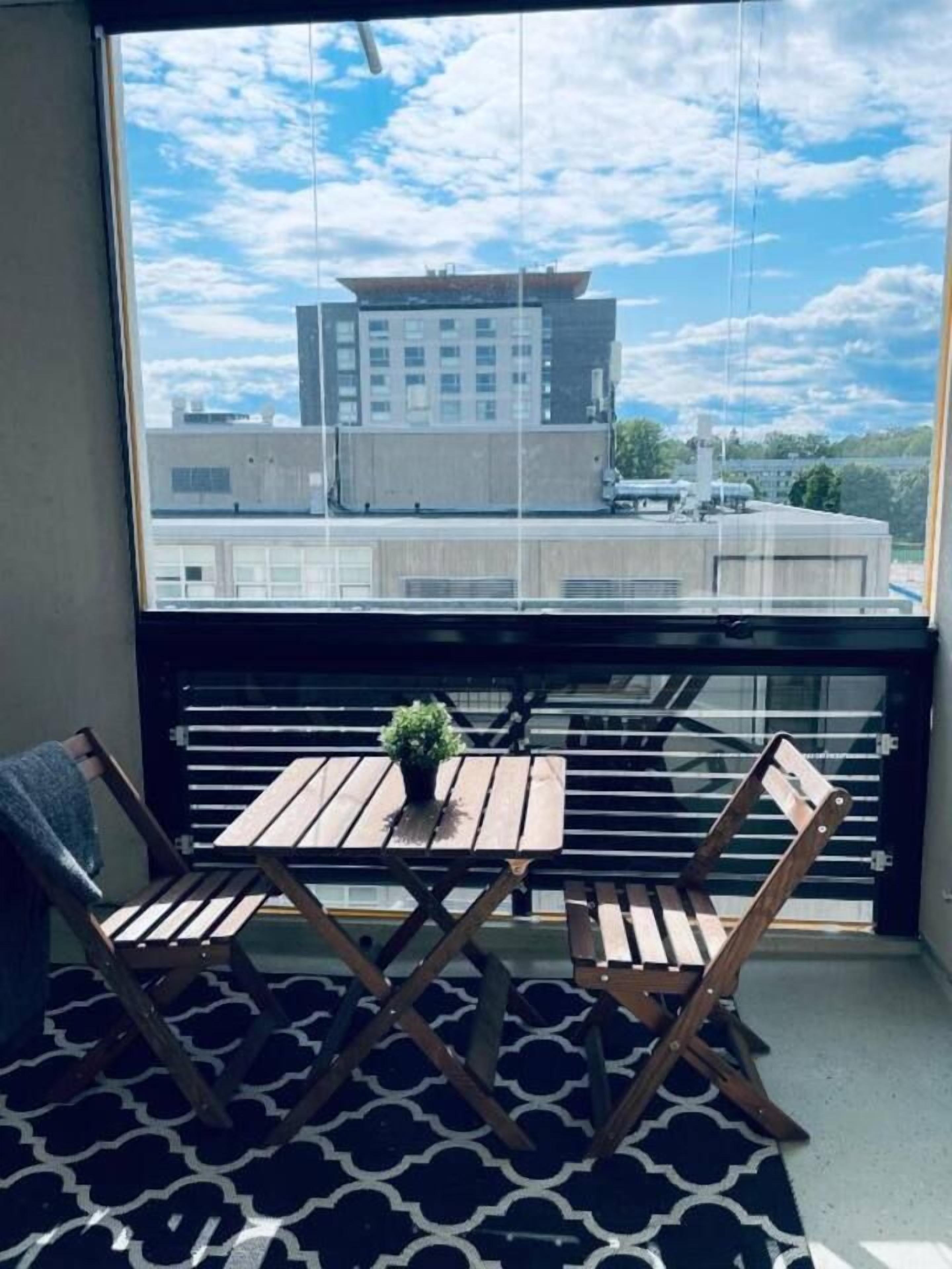 Un balcon avec une table et des chaises, un pot de fleurs et une vue sur les bâtiments à travers une fenêtre.