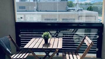 Un balcón con una mesa, sillas, una planta en maceta y vistas a edificios a través de una ventana.