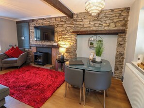 A cozy living room with a stone fireplace, a flat-screen TV, a red rug, and a dining area with a glass table. - Crown Cottage (Appleby-in-Westmorland)