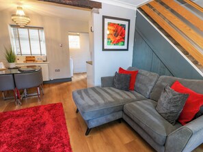 A living room with a grey sectional sofa, red and grey cushions, a red shag rug, a dining area with a glass table and chairs, a chandelier, and a staircase. - Crown Cottage (Appleby-in-Westmorland)