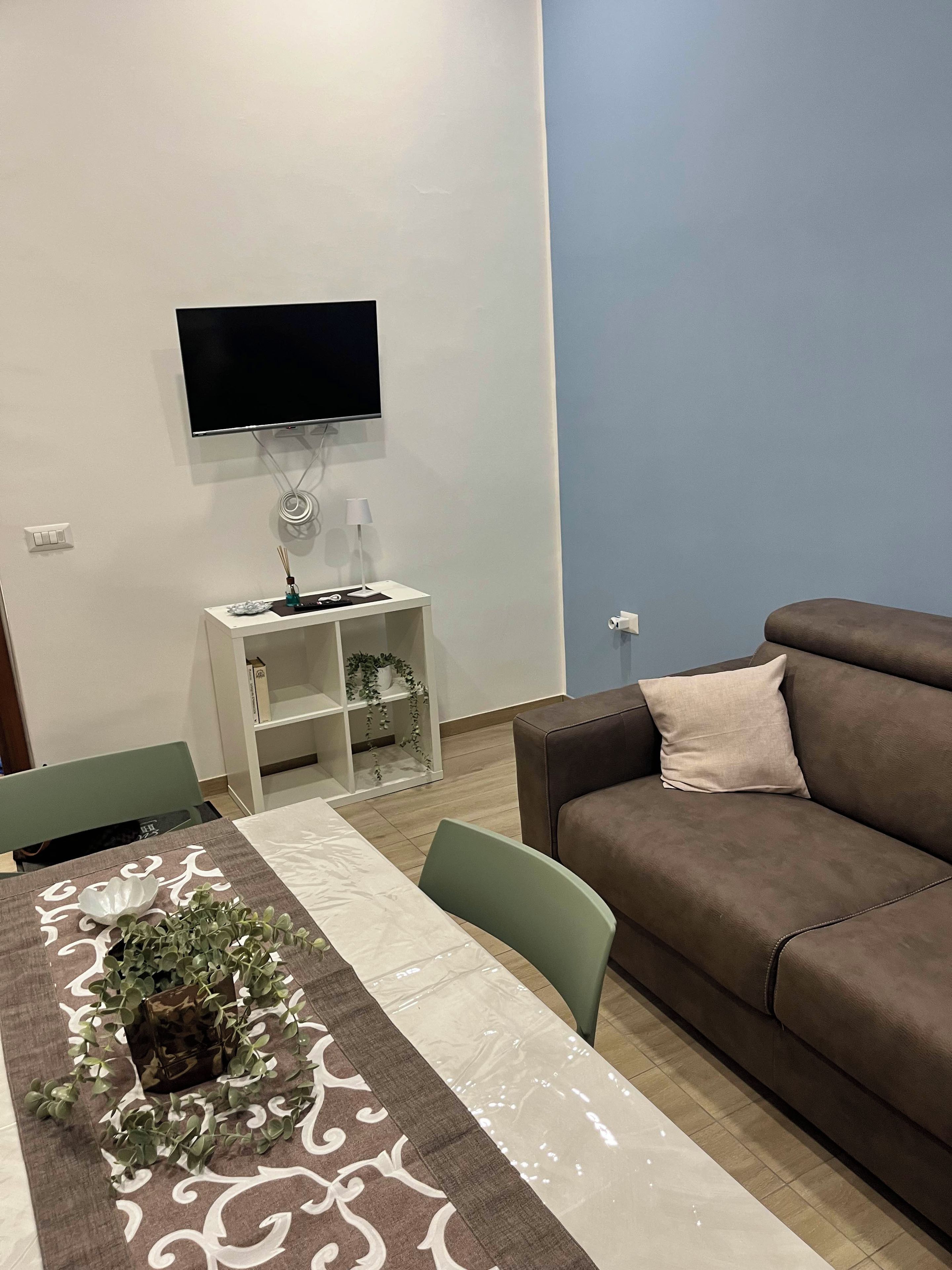 Ein Wohnzimmer mit braunem Sofa, einem an der Wand befestigten Fernseher, einem weißen Nachttisch mit Lampe und einem Esstisch mit gemustertem Tischläufer.