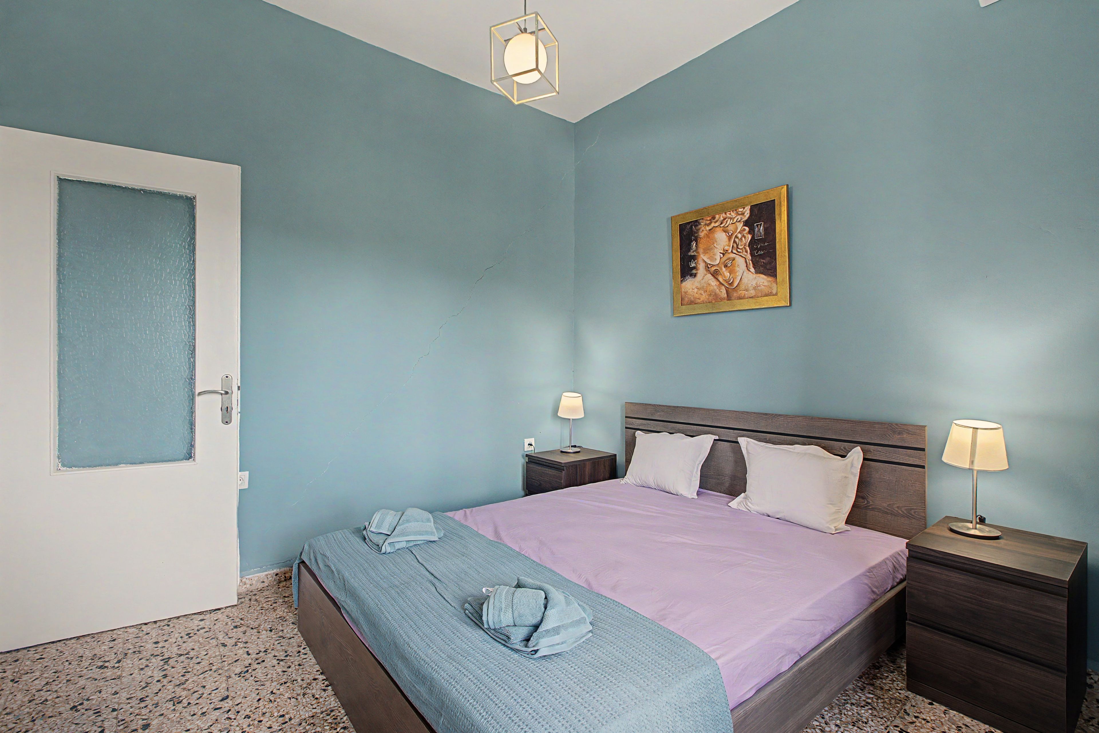 Un dormitorio con cama, dos mesitas de noche, una mesilla auxiliar, una puerta y un cuadro en la pared.