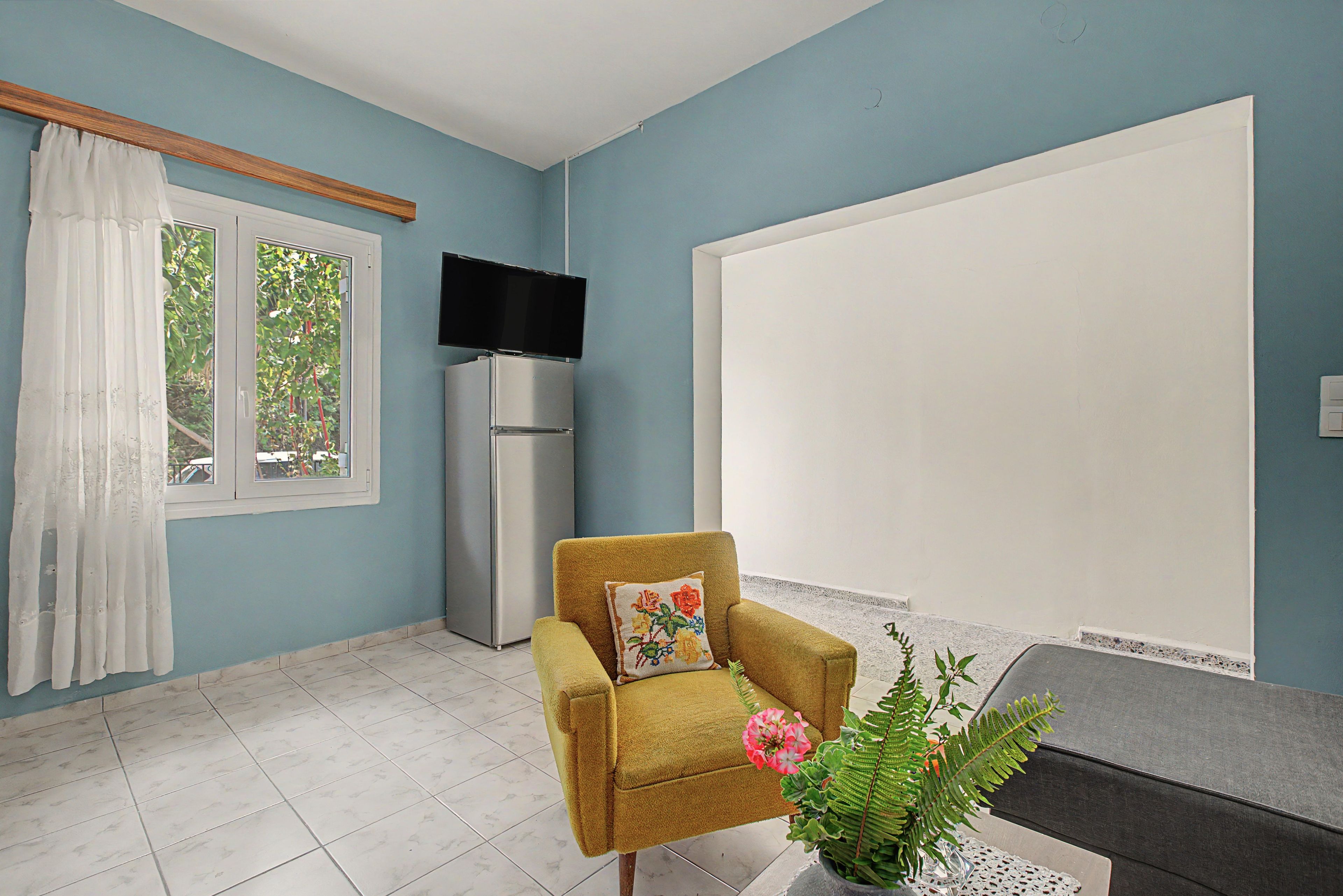 Habitación con sofá, televisión, refrigerador, ventana con cortinas y planta en maceta.