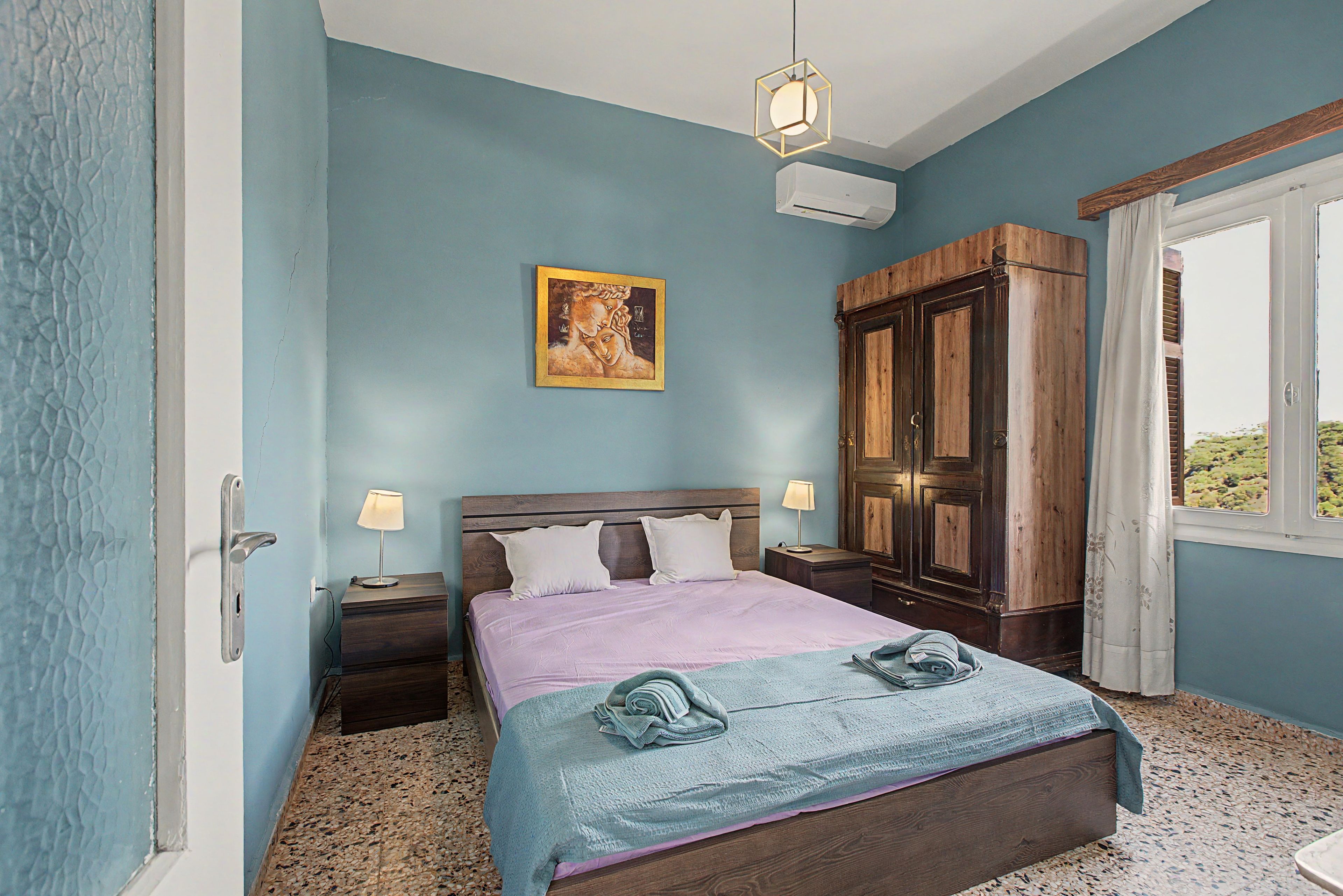 Une chambre à coucher avec un lit, deux tables de chevet, une commode, une armoire, un tableau au mur et une fenêtre.