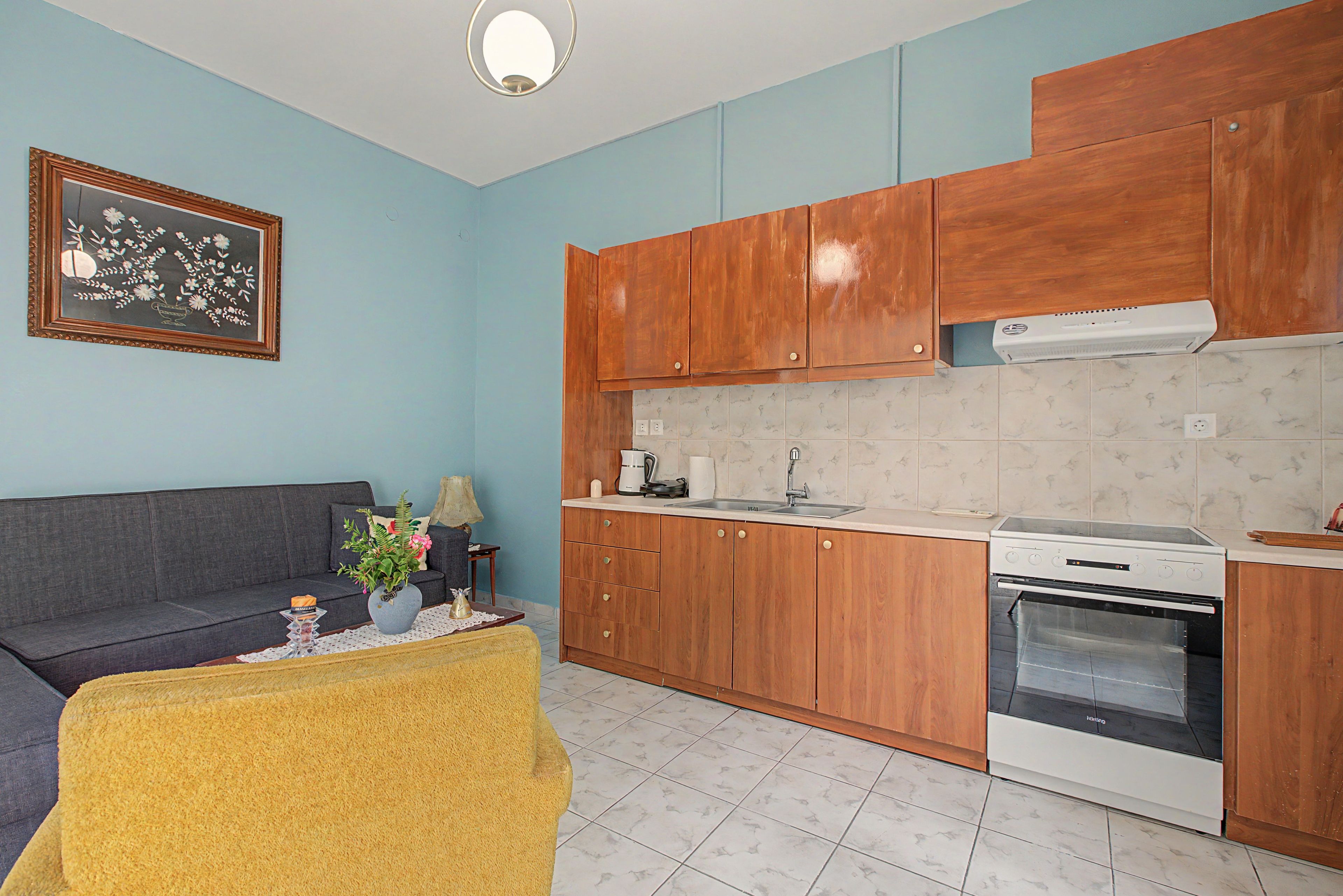 Een gezellig compact leefruimte met een kitchenette, een bank, een bijzettafeltje en een kunstwerk in een lijst aan de muur.