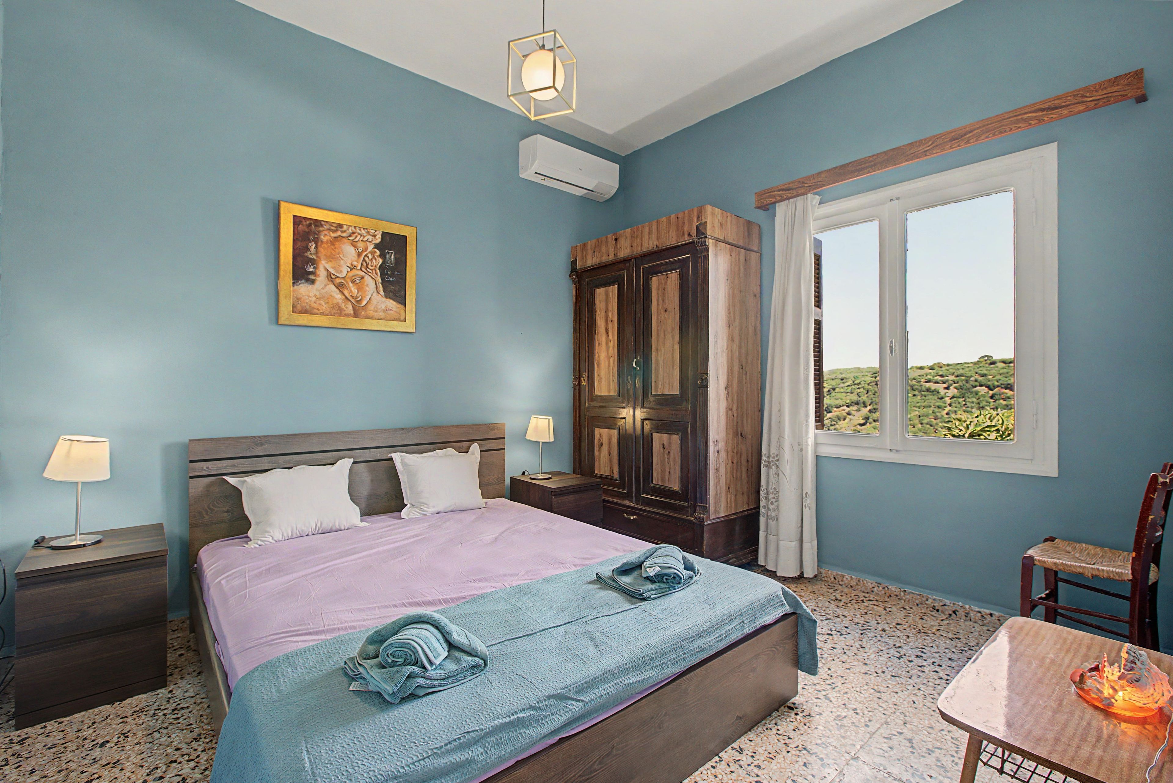 A bedroom with a bed, a nightstand, a lamp, a wardrobe, a window with a view, and a painting on the wall.