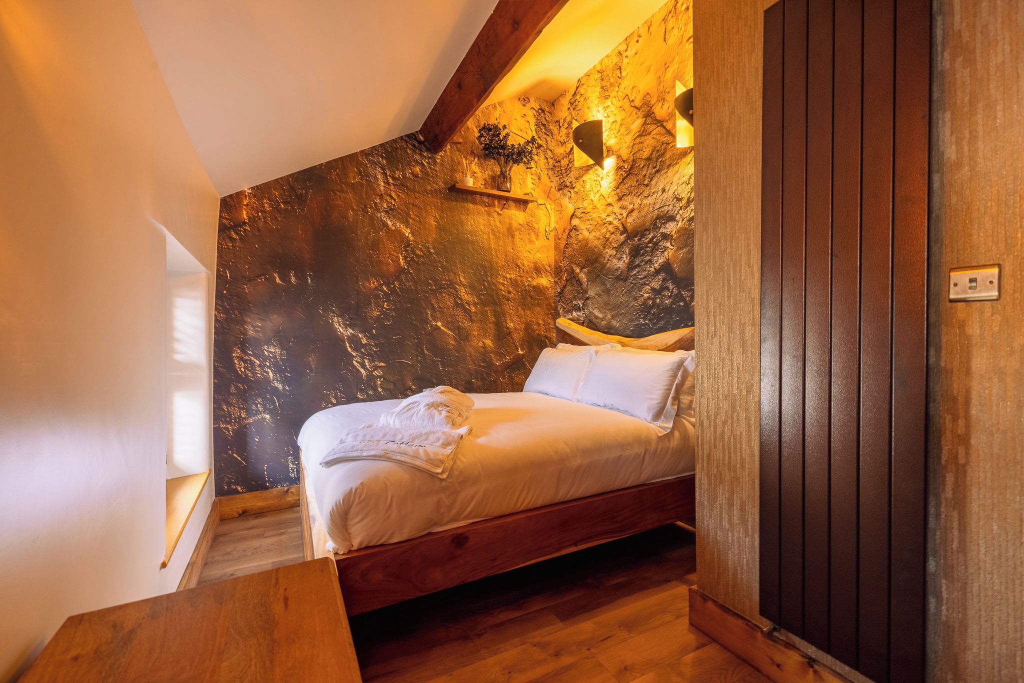 A bedroom with a wooden bed, a nightstand, a wall-mounted light, and a window.