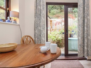 A dining area with a wooden table, chairs, and a view of a garden through a glass door. - The Cottage (Bridgwater)