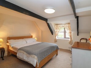 A bedroom with a wooden bed, a bedside table with a lamp, a window with curtains, and a desk with a chair. - The Cottage (Bridgwater)