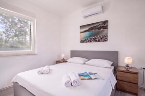 A neatly made bed with white linens and towels, a bedside table with a lamp, a wall-mounted air conditioner, and a framed picture of a coastal landscape. - Villa Blue Lagoon, Labin, Croatia (Labin)