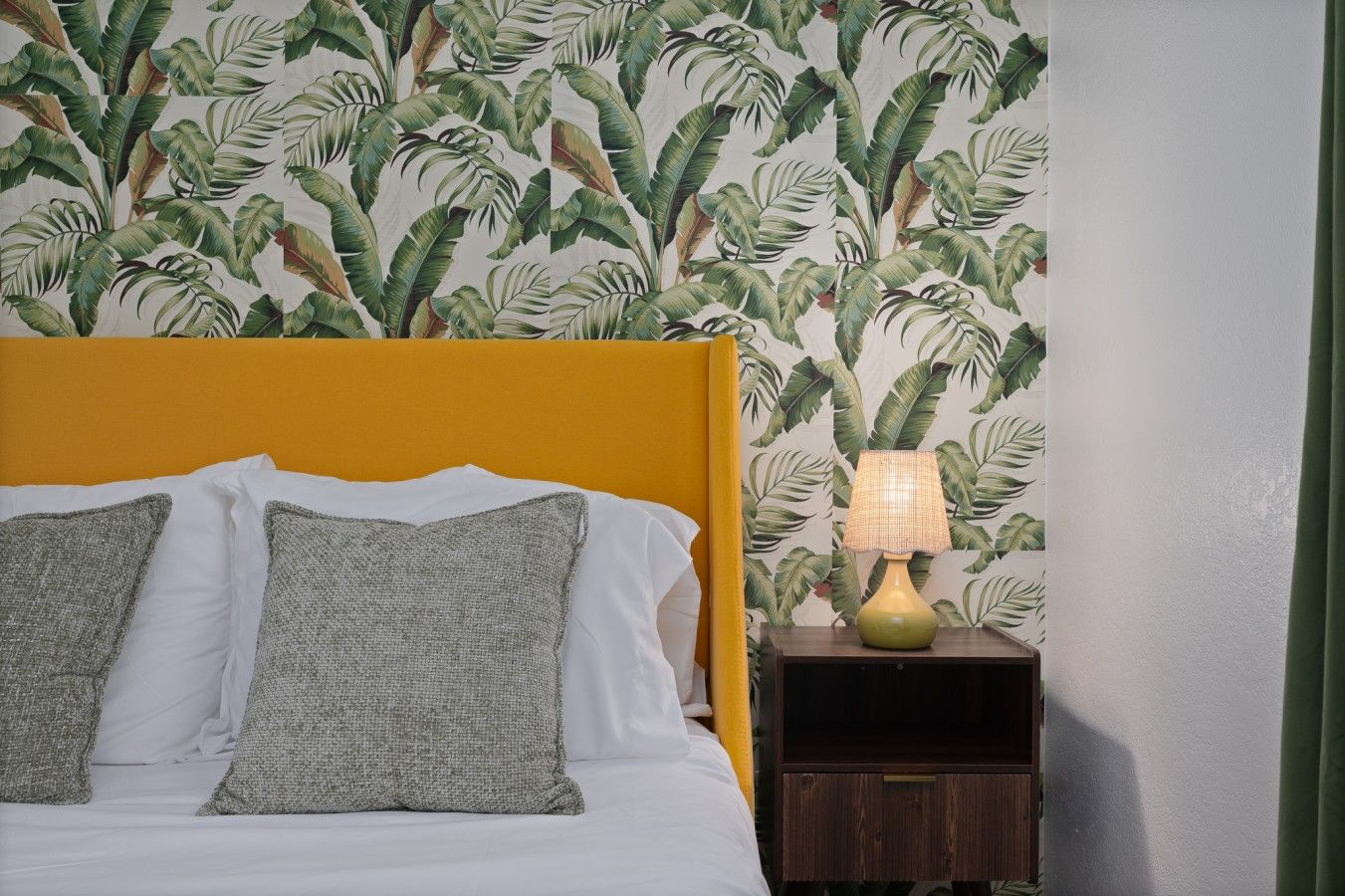 A bed with white linens and two gray pillows, a yellow headboard, and a nightstand with a lamp.