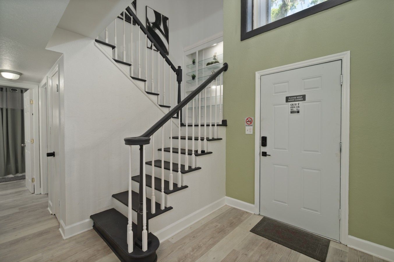 A staircase with black and white railings, a closed door with a 'Please Keep Door Closed' sign, and a doormat.