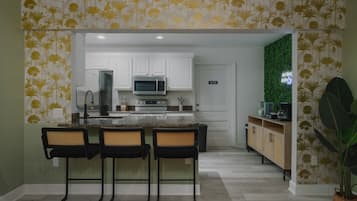 A kitchen with white cabinets, a microwave, and a sink. There are three black bar stools and a green plant.