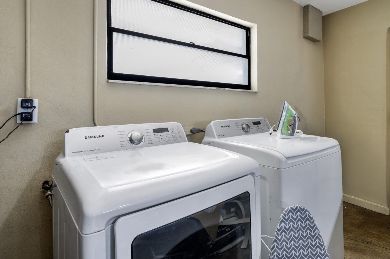 A laundry area with two Samsung washing machines, a window with blinds, and a wall-mounted air conditioner.