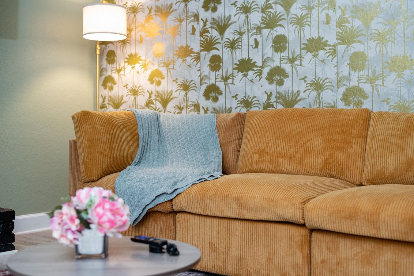 A living room with a brown sofa, a blue throw blanket, a lamp, a remote control, and a vase with flowers.