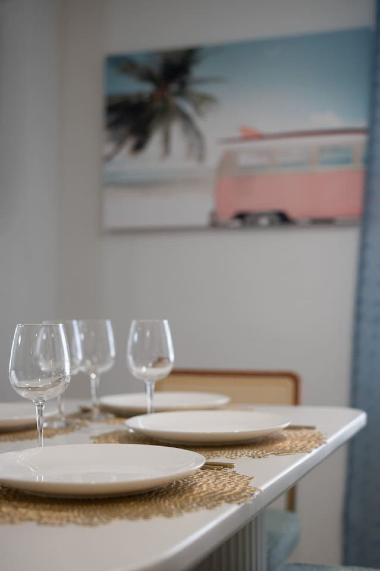 A dining table with plates and wine glasses, a woven placemat, and a painting of a pink car on the wall.