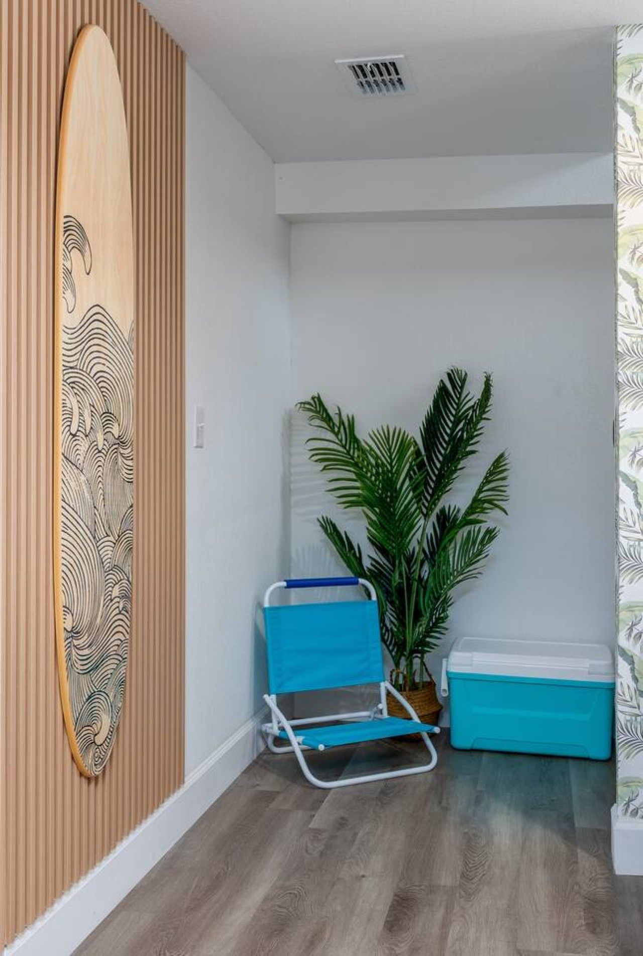Une pièce avec un mur lambrissé en bois, une décoration représentant une planche de surf, une chaise de plage bleue, une plante en pot et une glacière bleue.