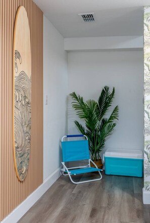 Et værelse med væg af træpaneler, en surfbræt-dekoration, en blå strandstol, en potteplante og en blå køleboks.