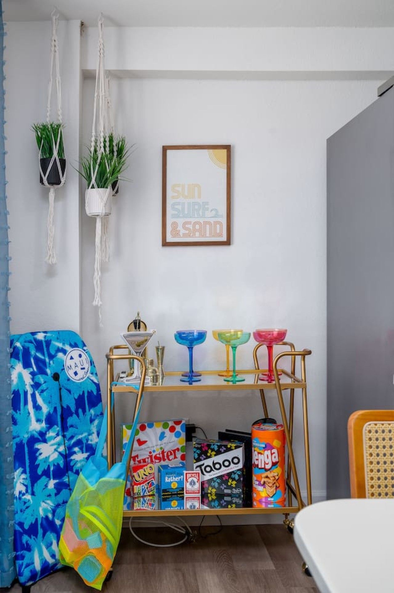 A room with a bed, a small table with colorful items, a macrame plant hanger, and a framed picture on the wall.
