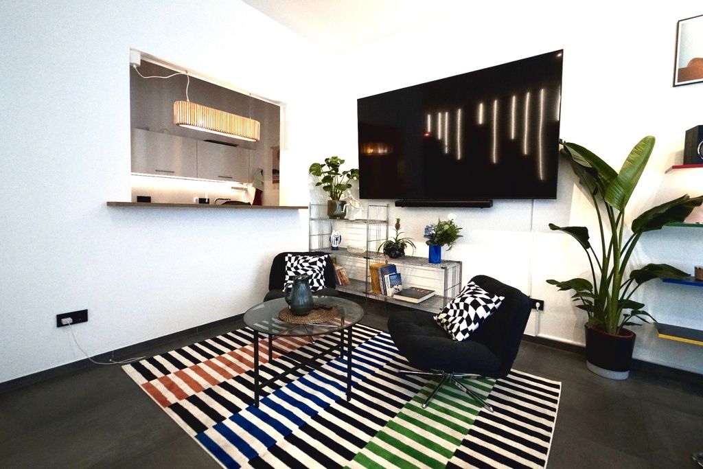 Ein modernes Wohnzimmer mit einem an der Wand befestigten Flachbildfernseher, einem Glastisch, zwei schwarzen Sesseln, einem Streifen Teppich und einer Topfpflanze.
