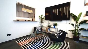 Ein modernes Wohnzimmer mit einem an der Wand befestigten Flachbildfernseher, einem Glastisch, zwei schwarzen Sesseln, einem Streifen Teppich und einer Topfpflanze.