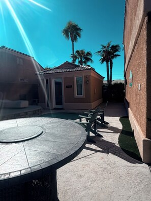Eine Terrasse mit einem runden Tisch und Stühlen, ein kleines Gebäude mit einer Tür und Palmen im Hintergrund.