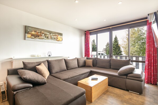 Ein modernes Wohnzimmer mit einem Ecksofa, einem Holztisch und einem großen Fenster mit Vorhängen.