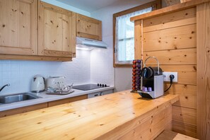 Una cocina con armarios de madera, salpicadero de azulejos blancos, una ventana con cortina blanca, una cafetera, un tostador y un enfriador de vino.