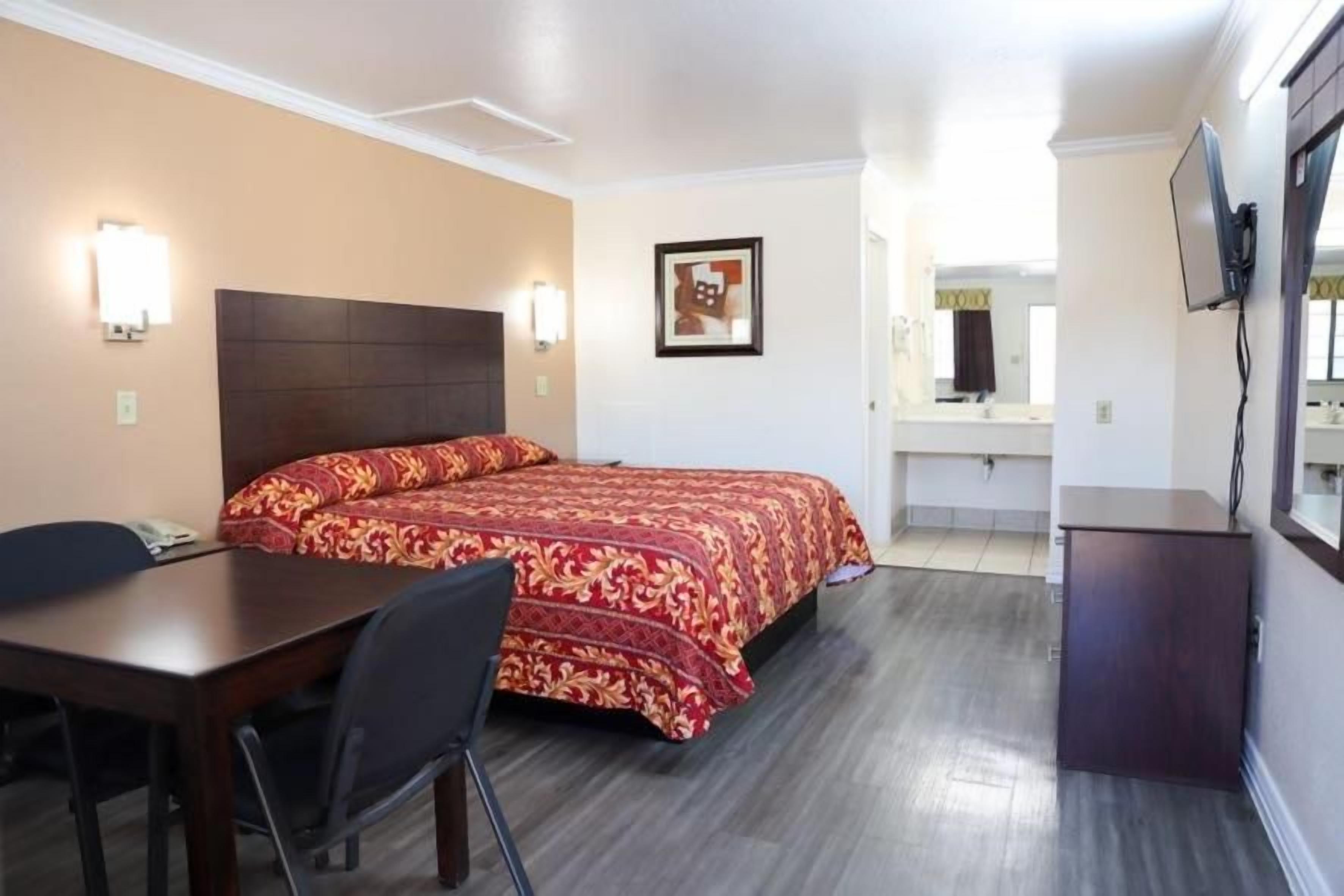 A hotel room with a bed, a desk, a chair, a TV, and a bathroom visible in the background.