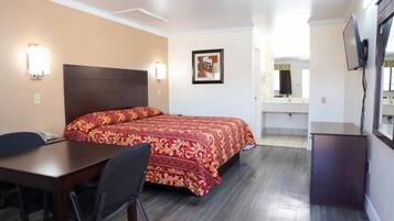 A hotel room with a bed, a desk, a chair, a TV, and a bathroom visible in the background.
