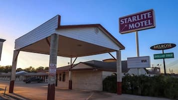 Um motel com área de estacionamento coberta, placa indicando que está aberto e uma placa próxima de uma loja Dollar Tree.