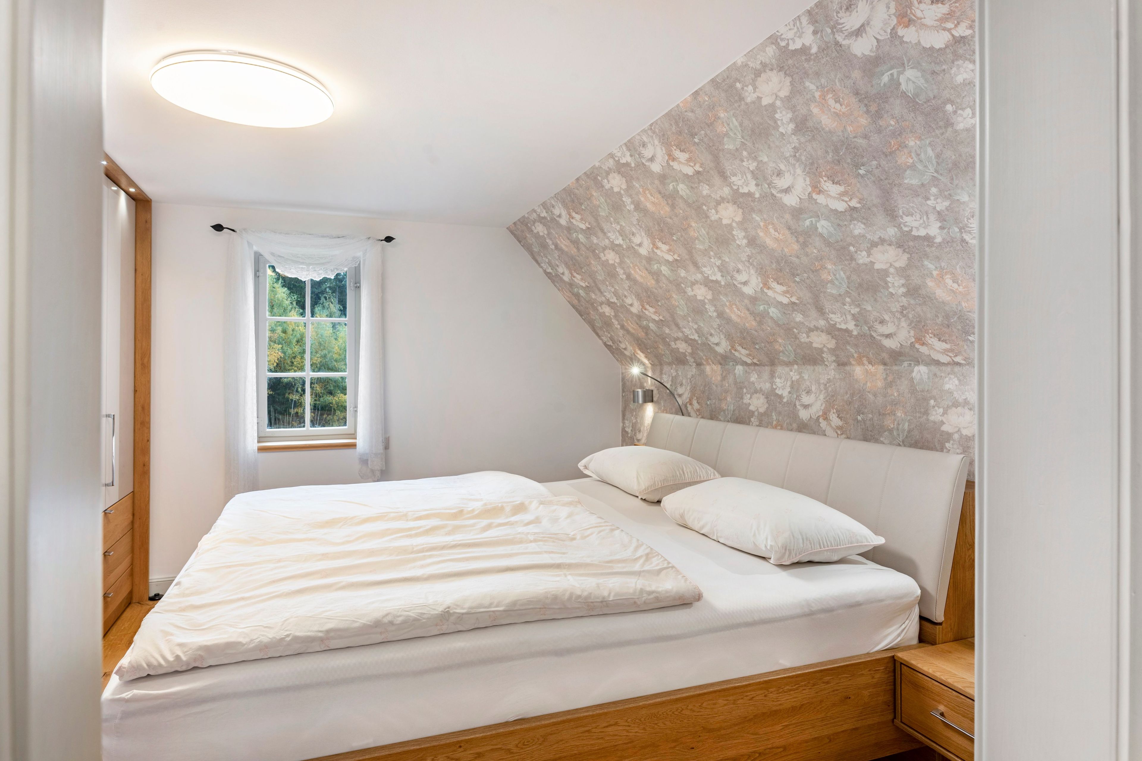A bedroom with a bed, a nightstand, a wall-mounted lamp, and a window with curtains.