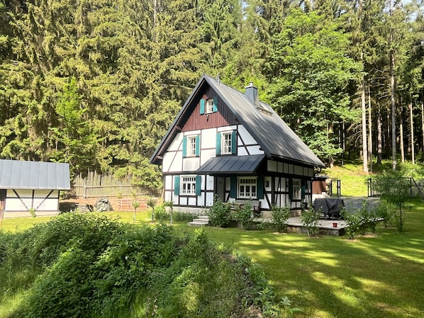 A traditional half-timbered house with a sloped roof, surrounded by a well-maintained garden and dense forest.