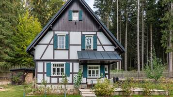 A traditional half-timbered house with a steep gable roof, surrounded by a forested area.