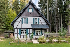 A traditional half-timbered house with a steep gable roof, surrounded by a forested area.