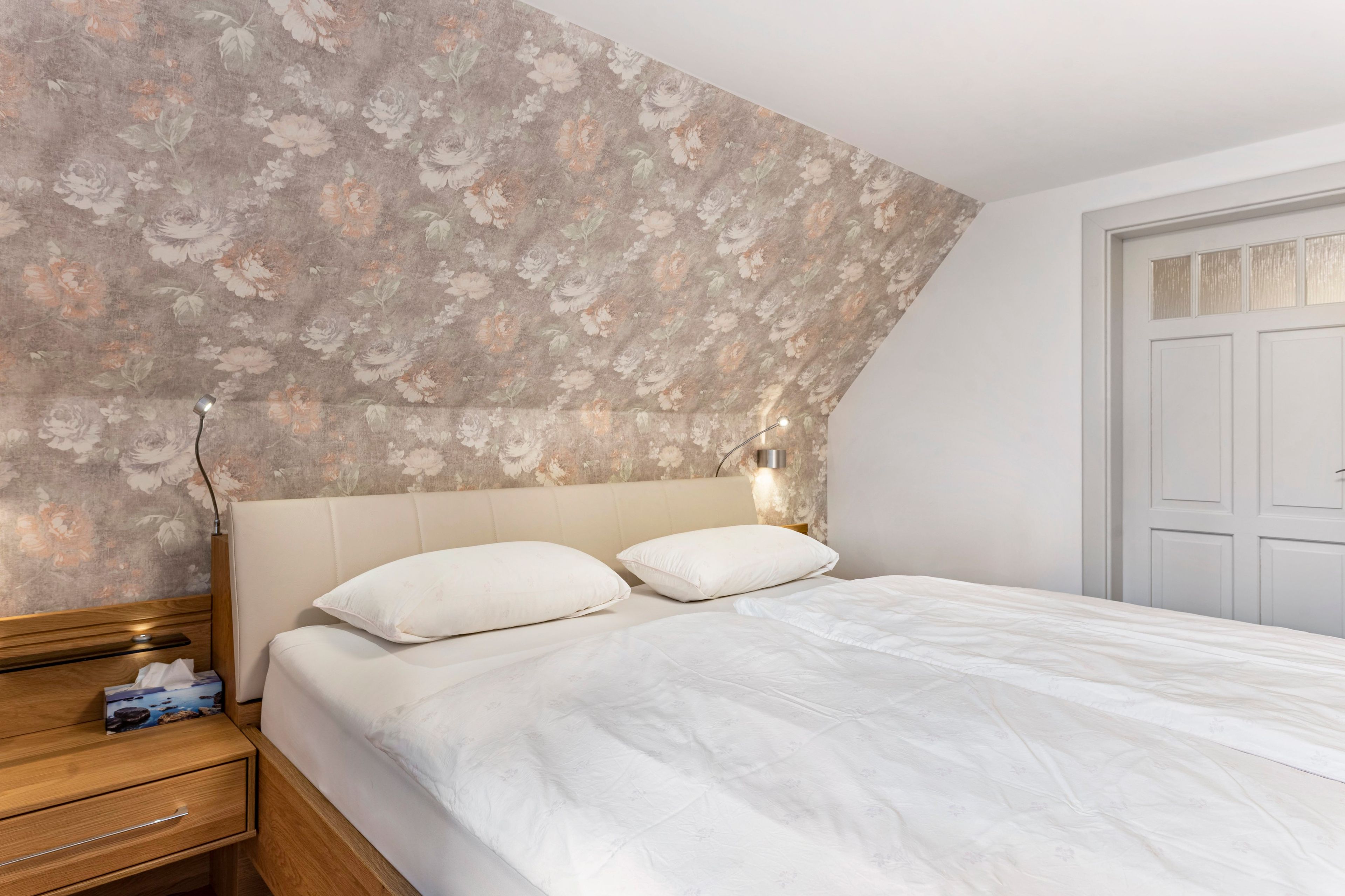 A bedroom with a floral wallpaper, a bed with white bedding, a nightstand with a lamp, and a wooden dresser.