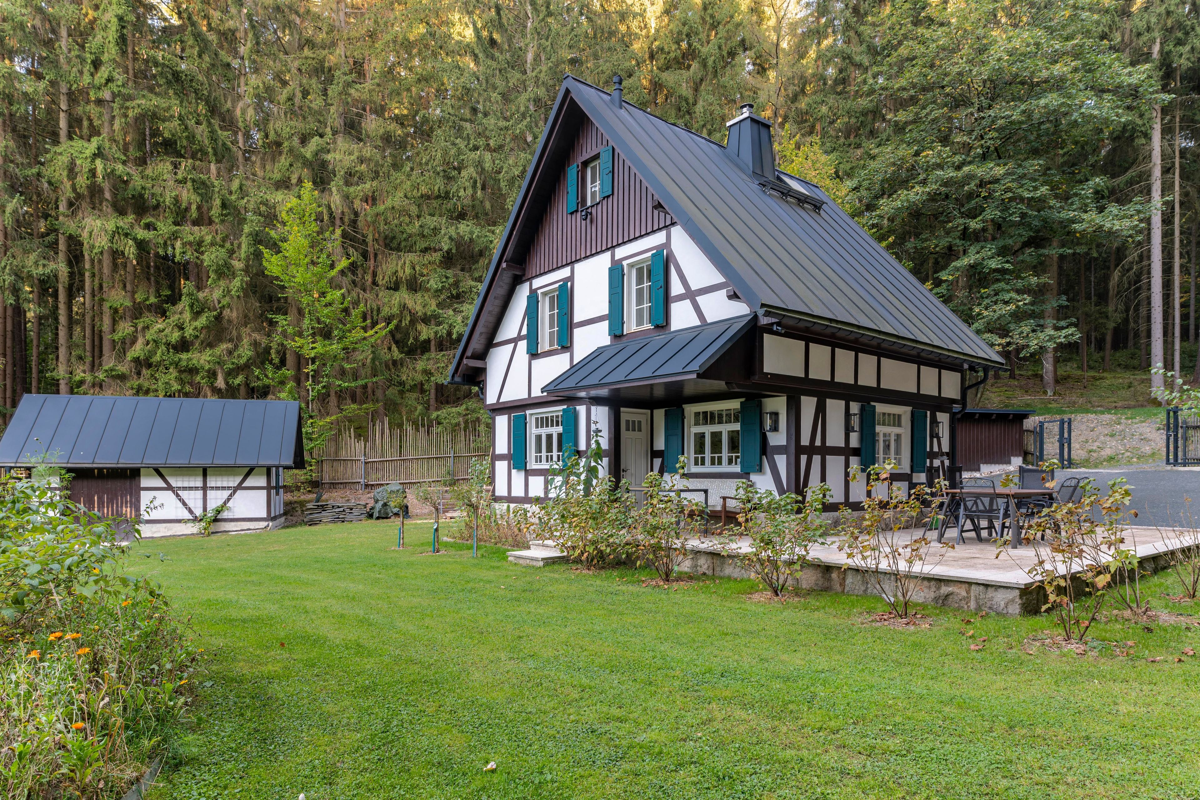 A traditional half-timbered house with a sloped roof, surrounded by a well-maintained lawn and a dense forest.
