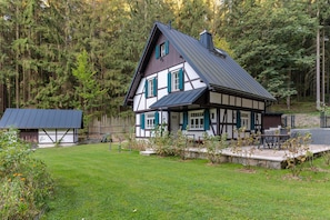 A traditional half-timbered house with a sloped roof, surrounded by a well-maintained lawn and a dense forest.