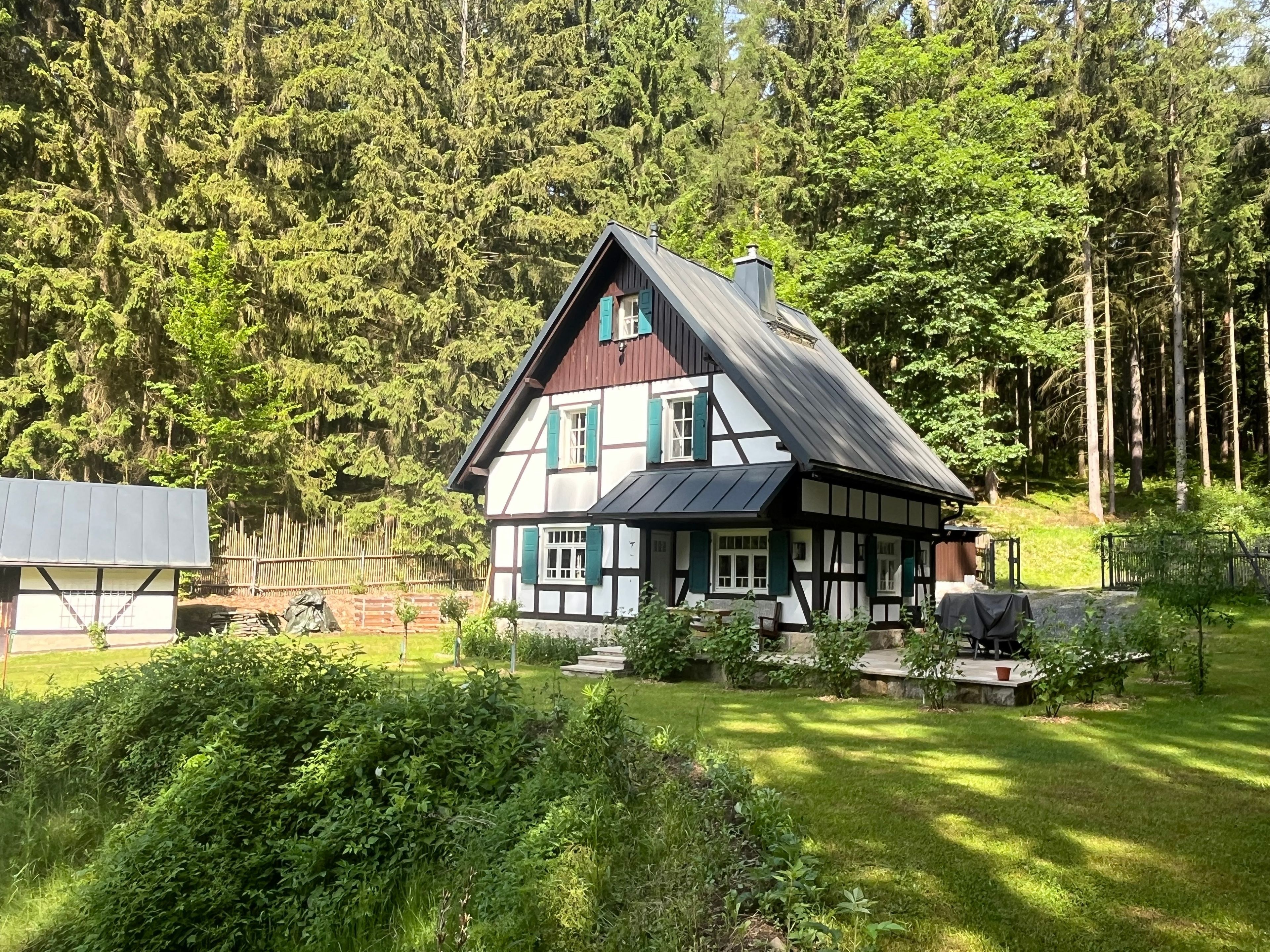 A traditional half-timbered house with a sloped roof, surrounded by a well-maintained garden and dense forest.