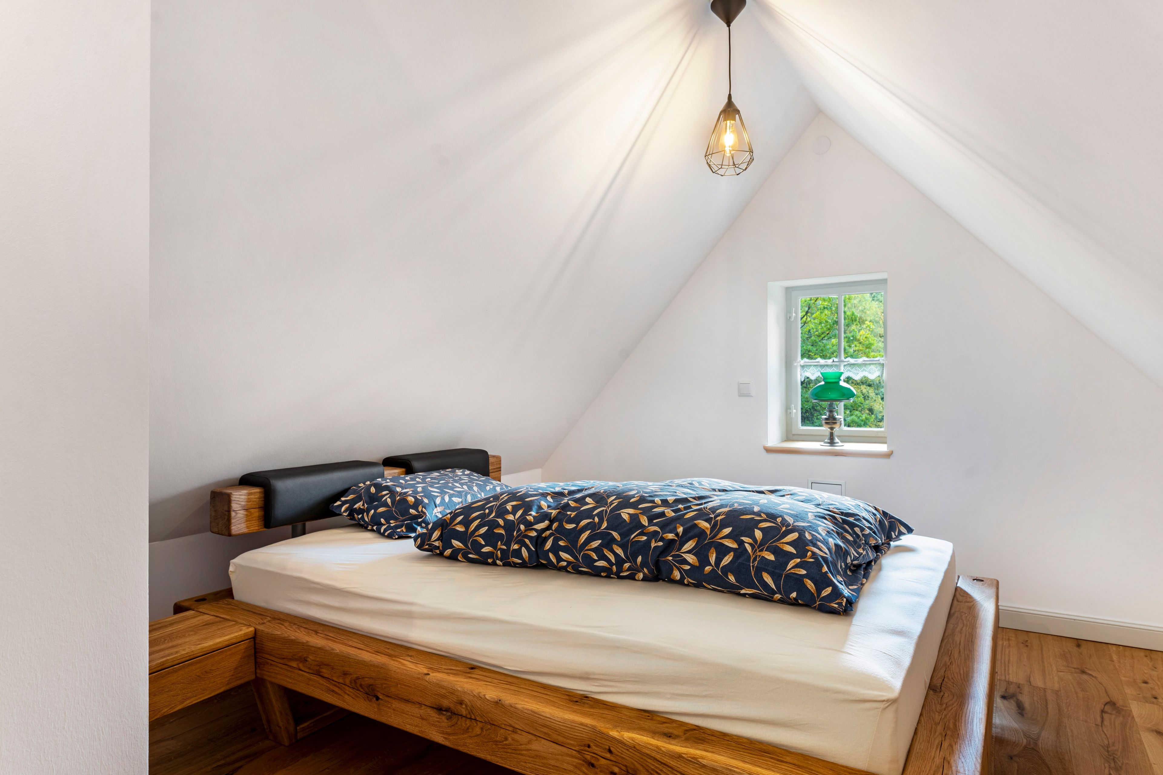 Un dormitorio con techo inclinado, cama con cabecera de madera, mesita de noche y ventana con vista a la vegetación.