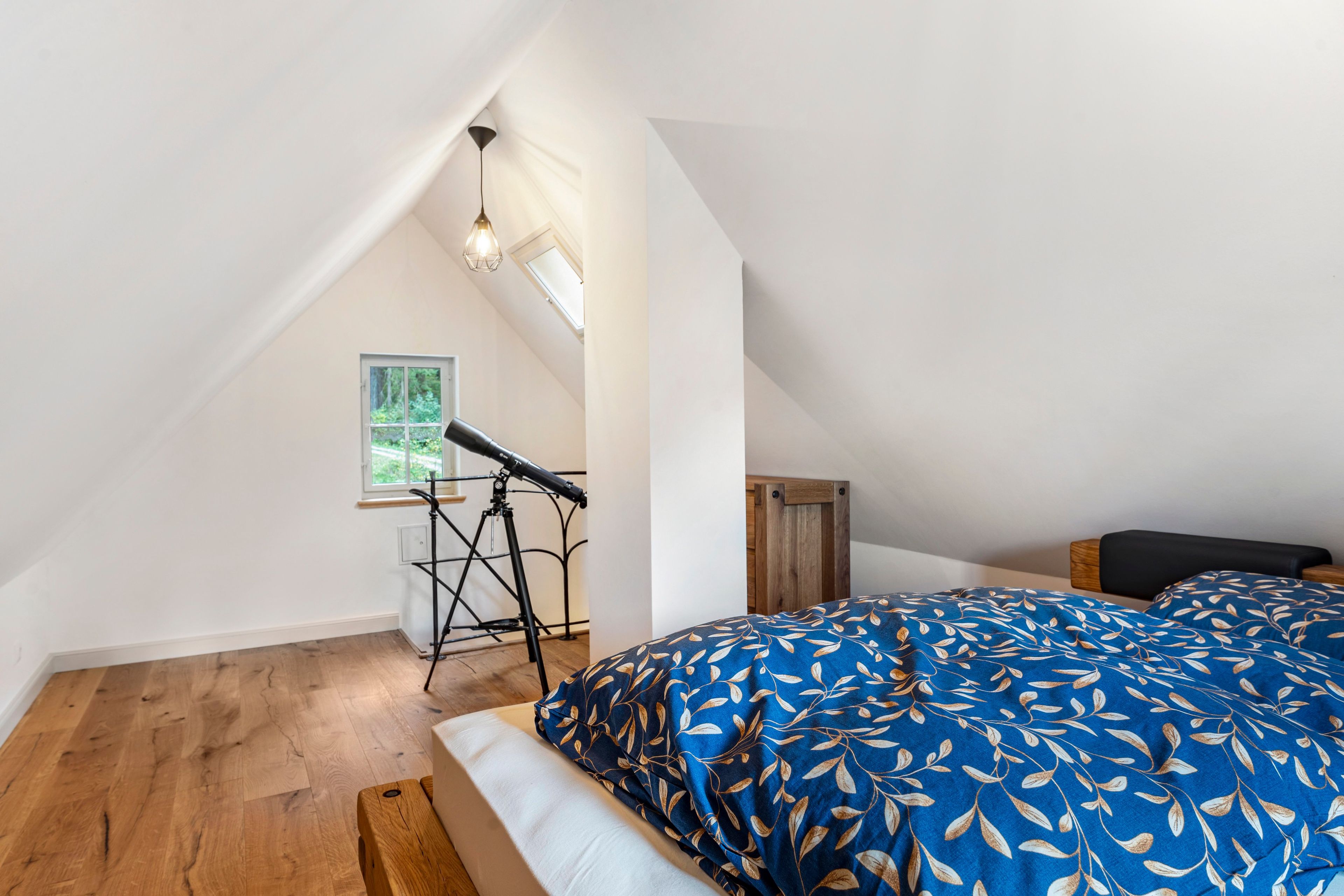 A bedroom with a sloped ceiling, a telescope, a bed with a blue leaf-patterned duvet, and a wooden bench.