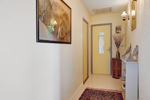 Un pasillo con una obra de arte enmarcadas, una puerta, un aplique de pared y una mesita con un florero y revistas.